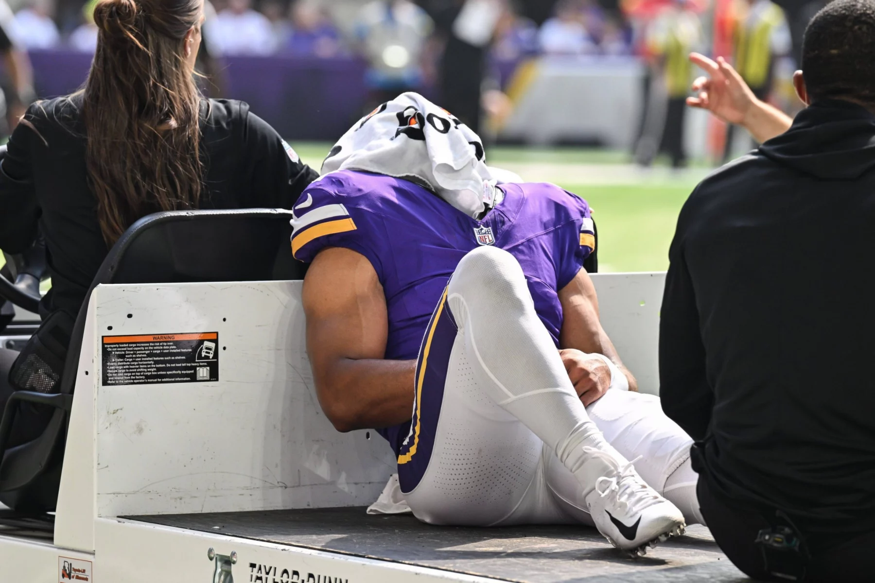 Ein Football-Spieler in einer lila Minnesota Vikings-Uniform liegt auf dem Rücken auf einem Krankenwagen, ein weißes Handtuch bedeckt seinen Kopf. Er scheint verletzt zu sein, ein Arm ruht auf seinem Knie - eine Erinnerung an die Momente, die Rondale Moore erlebt hat. Diese Beschreibung wurde mit der FootballR KI automatisch generiert.