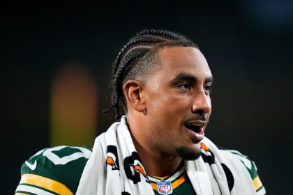 Ein Footballspieler, der Jordan Love mit geflochtenem Haar ähnelt, ein grün-gelbes Trikot trägt und ein schwarz-weißes Handtuch um die Schultern geschlungen hat, schaut mit offenem Mund zur Seite. Der dunkle, unscharfe Hintergrund lenkt die Aufmerksamkeit auf sein Gesicht und seine Uniform. Diese Beschreibung wurde mit der FootballR KI automatisch generiert.