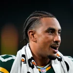 NFL Spieltag 2 - Week 4 - Ein Footballspieler, der Jordan Love mit geflochtenem Haar ähnelt, ein grün-gelbes Trikot trägt und ein schwarz-weißes Handtuch um die Schultern geschlungen hat, schaut mit offenem Mund zur Seite. Der dunkle, unscharfe Hintergrund lenkt die Aufmerksamkeit auf sein Gesicht und seine Uniform. Diese Beschreibung wurde mit der FootballR KI automatisch generiert.