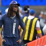 Der Star der Dallas Cowboys, CeeDee Lamb, mit Sonnenbrille, marineblauem Hemd und "D"-Logo-Mütze, steht mit langen Dreadlocks und mehrlagigen Ketten an der Seitenlinie. Andere Teammitglieder und Mitarbeiter in blauen und gelben Westen sind im unscharfen Hintergrund zu sehen. Diese Beschreibung wurde mit der FootballR KI automatisch generiert.