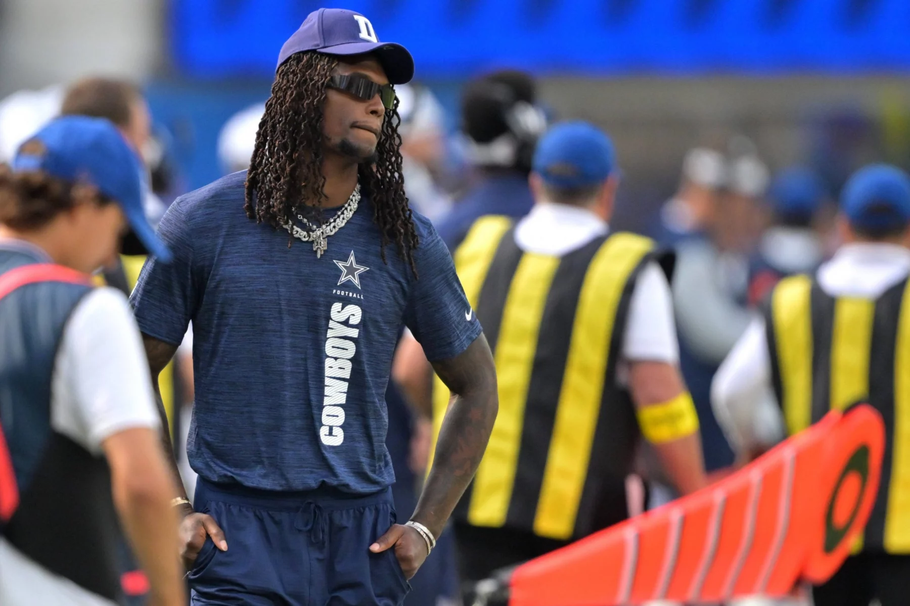 Der Star der Dallas Cowboys, CeeDee Lamb, mit Sonnenbrille, marineblauem Hemd und "D"-Logo-Mütze, steht mit langen Dreadlocks und mehrlagigen Ketten an der Seitenlinie. Andere Teammitglieder und Mitarbeiter in blauen und gelben Westen sind im unscharfen Hintergrund zu sehen. Diese Beschreibung wurde mit der FootballR KI automatisch generiert.