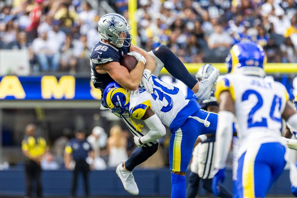 Ein Footballspieler in einem blauen Trikot der Dallas Cowboys wird während der NFL Preseason von einem Verteidiger der Los Angeles Rams in der Luft angegriffen. Der Cowboys-Spieler hält den Ball fest umklammert, als der Rams-Spieler ihn vom überfüllten Feld hebt. Diese Beschreibung wurde mit der FootballR KI automatisch generiert.