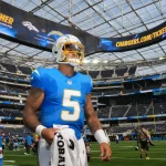 Ein Footballspieler, Trey Lance, in einem blauen Los Angeles Chargers Trikot und Helm, Nummer 5, steht auf dem Spielfeld im SoFi Stadium. Er hält ein weißes Handtuch in der Hand. Unter der großen Anzeigetafel für die Heimpremiere der Chargers sind Teamkollegen und Fans zu sehen. Diese Beschreibung wurde mit der FootballR KI automatisch generiert.