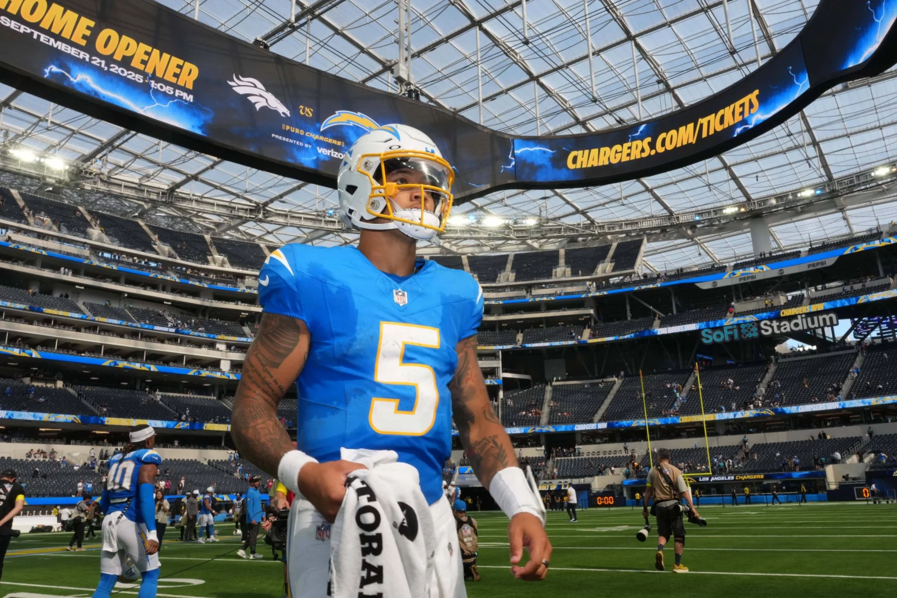 Ein Footballspieler, Trey Lance, in einem blauen Los Angeles Chargers Trikot und Helm, Nummer 5, steht auf dem Spielfeld im SoFi Stadium. Er hält ein weißes Handtuch in der Hand. Unter der großen Anzeigetafel für die Heimpremiere der Chargers sind Teamkollegen und Fans zu sehen. Diese Beschreibung wurde mit der FootballR KI automatisch generiert.