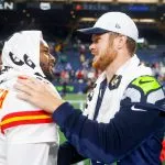 Zwei NFL-Spieler gegnerischer Mannschaften, einer in der Uniform der Kansas City Chiefs, der andere in der Uniform der Seattle Seahawks, lächeln und geben sich nach einem NFL Preseason Spiel die Hand. Beide haben Handtücher über die Schultern geworfen, während im Hintergrund die Stadionbeleuchtung und die Fans zu sehen sind. Diese Beschreibung wurde mit der FootballR KI automatisch generiert.