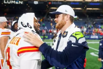 Zwei NFL-Spieler gegnerischer Mannschaften, einer in der Uniform der Kansas City Chiefs, der andere in der Uniform der Seattle Seahawks, lächeln und geben sich nach einem NFL Preseason Spiel die Hand. Beide haben Handtücher über die Schultern geworfen, während im Hintergrund die Stadionbeleuchtung und die Fans zu sehen sind. Diese Beschreibung wurde mit der FootballR KI automatisch generiert.