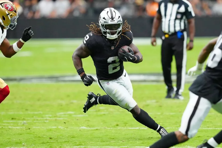 Ein Footballspieler in einem schwarzen Trikot der Las Vegas Raiders, der mit seinen langen Haaren Ashton Jeanty ähnelt, läuft während eines Spiels mit dem Ball. Er wird von Spielern der San Francisco 49ers auf einem grünen Feld verfolgt, im Hintergrund ist ein Offizieller zu sehen. Diese Beschreibung wurde mit der FootballR KI automatisch generiert.