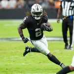 Ein Footballspieler in einem schwarzen Trikot der Las Vegas Raiders, der mit seinen langen Haaren Ashton Jeanty ähnelt, läuft während eines Spiels mit dem Ball. Er wird von Spielern der San Francisco 49ers auf einem grünen Feld verfolgt, im Hintergrund ist ein Offizieller zu sehen. Diese Beschreibung wurde mit der FootballR KI automatisch generiert.