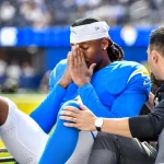 Quentin Johnston in einem blau-gelben Trikot sitzt auf einem Wagen, hat die Augen geschlossen und die Hände vor das Gesicht gehalten. Er wirkt aufgewühlt oder schmerzerfüllt - möglicherweise hat er eine Gehirnerschütterung erlitten -, während ein Trainer oder Betreuer ihm Trost spendet, indem er seine Schulter und sein Knie berührt. Diese Beschreibung wurde mit der FootballR KI automatisch generiert.