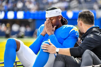 Quentin Johnston in einem blau-gelben Trikot sitzt auf einem Wagen, hat die Augen geschlossen und die Hände vor das Gesicht gehalten. Er wirkt aufgewühlt oder schmerzerfüllt - möglicherweise hat er eine Gehirnerschütterung erlitten -, während ein Trainer oder Betreuer ihm Trost spendet, indem er seine Schulter und sein Knie berührt. Diese Beschreibung wurde mit der FootballR KI automatisch generiert.