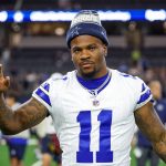 Micah Parsons, in einem weißen Dallas Cowboys-Trikot mit der Nummer 11 und einer blauen Mütze, steht auf dem Spielfeld und schaut in die Kamera, einen Arm leicht erhoben. Im verschwommenen Stadionhintergrund sind andere Spieler und helle Lichter zu sehen. Diese Beschreibung wurde mit der FootballR KI automatisch generiert.