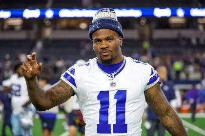 Micah Parsons, in einem weißen Dallas Cowboys-Trikot mit der Nummer 11 und einer blauen Mütze, steht auf dem Spielfeld und schaut in die Kamera, einen Arm leicht erhoben. Im verschwommenen Stadionhintergrund sind andere Spieler und helle Lichter zu sehen. Diese Beschreibung wurde mit der FootballR KI automatisch generiert.
