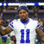 Micah Parsons, in einem weißen Dallas Cowboys-Trikot mit der Nummer 11 und einer blauen Mütze, steht auf dem Spielfeld und schaut in die Kamera, einen Arm leicht erhoben. Im verschwommenen Stadionhintergrund sind andere Spieler und helle Lichter zu sehen. Diese Beschreibung wurde mit der FootballR KI automatisch generiert.
