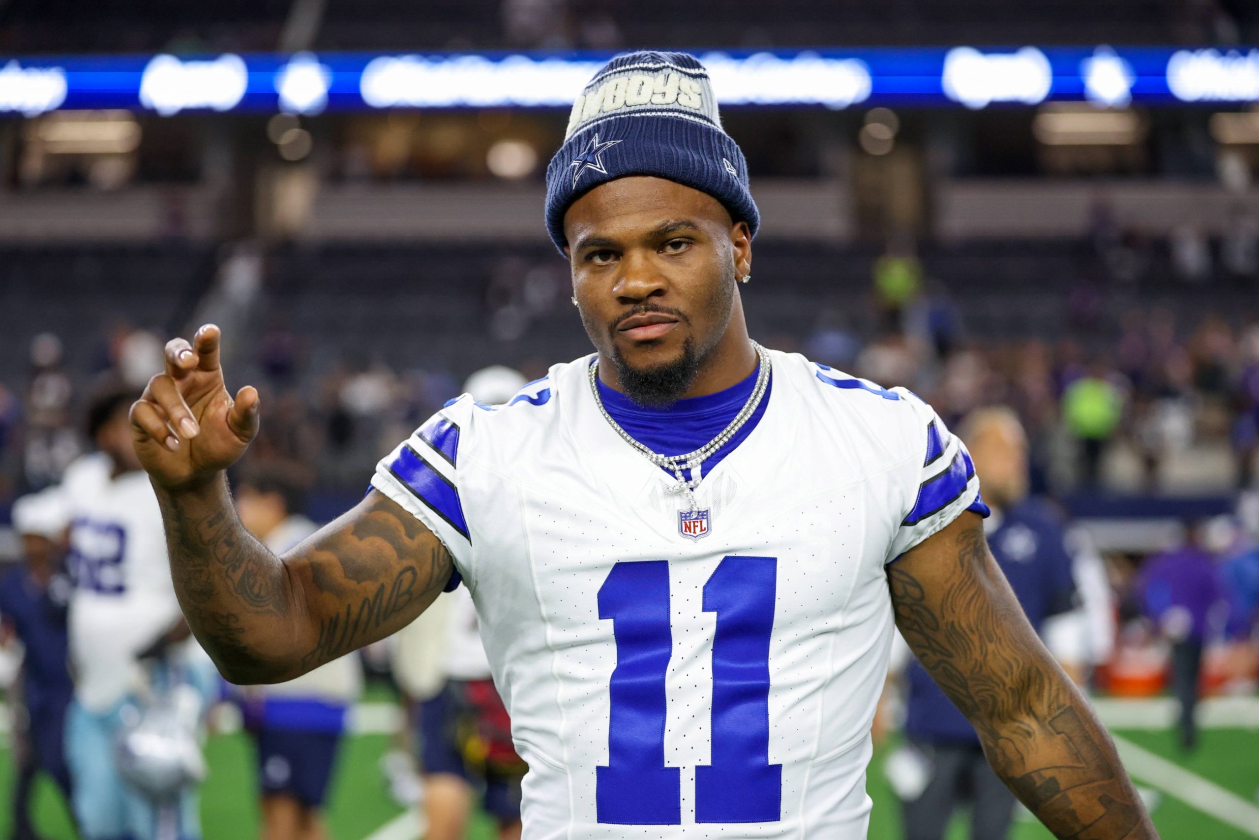 Micah Parsons, in einem weißen Dallas Cowboys-Trikot mit der Nummer 11 und einer blauen Mütze, steht auf dem Spielfeld und schaut in die Kamera, einen Arm leicht erhoben. Im verschwommenen Stadionhintergrund sind andere Spieler und helle Lichter zu sehen. Diese Beschreibung wurde mit der FootballR KI automatisch generiert.
