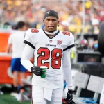 Ein Footballspieler, Shilo Sanders, trägt ein weißes Trikot der Tampa Bay Buccaneers mit der Nummer 28 und hält einen grünen Becher an der Seitenlinie. Sein Helm ist abgenommen, und er hat einen konzentrierten Gesichtsausdruck. Im Hintergrund sind andere Spieler und eine unscharfe Menge zu sehen. Diese Beschreibung wurde mit der FootballR KI automatisch generiert.