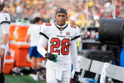 Ein Footballspieler, Shilo Sanders, trägt ein weißes Trikot der Tampa Bay Buccaneers mit der Nummer 28 und hält einen grünen Becher an der Seitenlinie. Sein Helm ist abgenommen, und er hat einen konzentrierten Gesichtsausdruck. Im Hintergrund sind andere Spieler und eine unscharfe Menge zu sehen. Diese Beschreibung wurde mit der FootballR KI automatisch generiert.