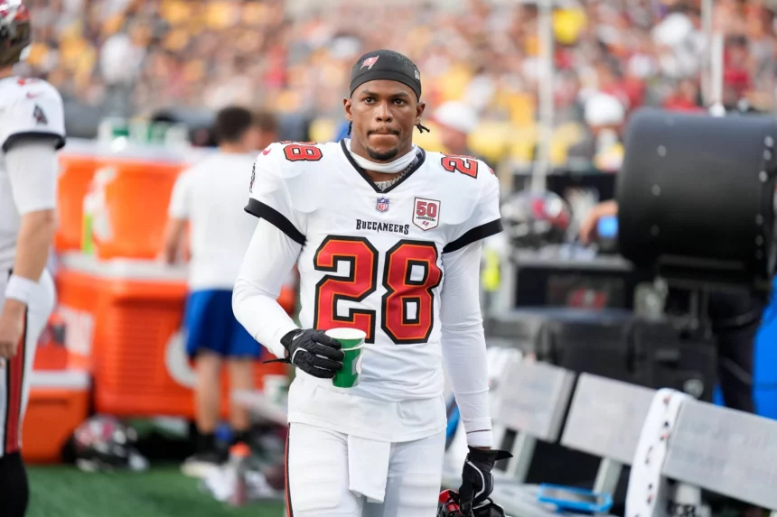 Ein Footballspieler, Shilo Sanders, trägt ein weißes Trikot der Tampa Bay Buccaneers mit der Nummer 28 und hält einen grünen Becher an der Seitenlinie. Sein Helm ist abgenommen, und er hat einen konzentrierten Gesichtsausdruck. Im Hintergrund sind andere Spieler und eine unscharfe Menge zu sehen. Diese Beschreibung wurde mit der FootballR KI automatisch generiert.