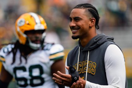 Ein lächelnder Jordan Love in einem grauen Packers-Kapuzenpulli klatscht an der Seitenlinie in die Hände. Er trägt kurze Zöpfe und ein schwarzes Armband, während ein anderer Packers-Spieler in Uniform und Helm unscharf im Hintergrund auf dem Football-Feld steht. Diese Beschreibung wurde mit der FootballR KI automatisch generiert.