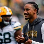 Ein lächelnder Jordan Love in einem grauen Packers-Kapuzenpulli klatscht an der Seitenlinie in die Hände. Er trägt kurze Zöpfe und ein schwarzes Armband, während ein anderer Packers-Spieler in Uniform und Helm unscharf im Hintergrund auf dem Football-Feld steht. Diese Beschreibung wurde mit der FootballR KI automatisch generiert.