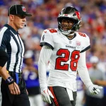 Ein Footballspieler der Tampa Bay Buccaneers, der Shilo Sanders ähnelt und die Trikotnummer 28 trägt, steht auf dem Spielfeld neben einem NFL-Schiedsrichter. Der Spieler, der eine weiße Uniform mit schwarzen und roten Akzenten trägt, blickt zur Seite, während eine unscharfe Menschenmenge den Hintergrund füllt. Diese Beschreibung wurde mit der FootballR KI automatisch generiert.