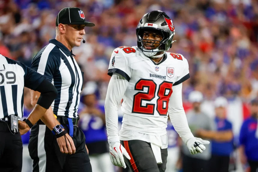 Ein Footballspieler der Tampa Bay Buccaneers, der Shilo Sanders ähnelt und die Trikotnummer 28 trägt, steht auf dem Spielfeld neben einem NFL-Schiedsrichter. Der Spieler, der eine weiße Uniform mit schwarzen und roten Akzenten trägt, blickt zur Seite, während eine unscharfe Menschenmenge den Hintergrund füllt. Diese Beschreibung wurde mit der FootballR KI automatisch generiert.