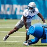 Morice Norris, ein Footballspieler in Trainingskleidung der Detroit Lions mit silbernem Helm und blauen Handschuhen, beugt sich auf dem Rasen vor, um einen blauen Tackling-Dummy zu schieben. Eine weitere Person in schwarzer Sportkleidung steht in der Nähe, im verschwommenen Hintergrund ist "LIONS" zu sehen. Diese Beschreibung wurde mit der FootballR KI automatisch generiert.