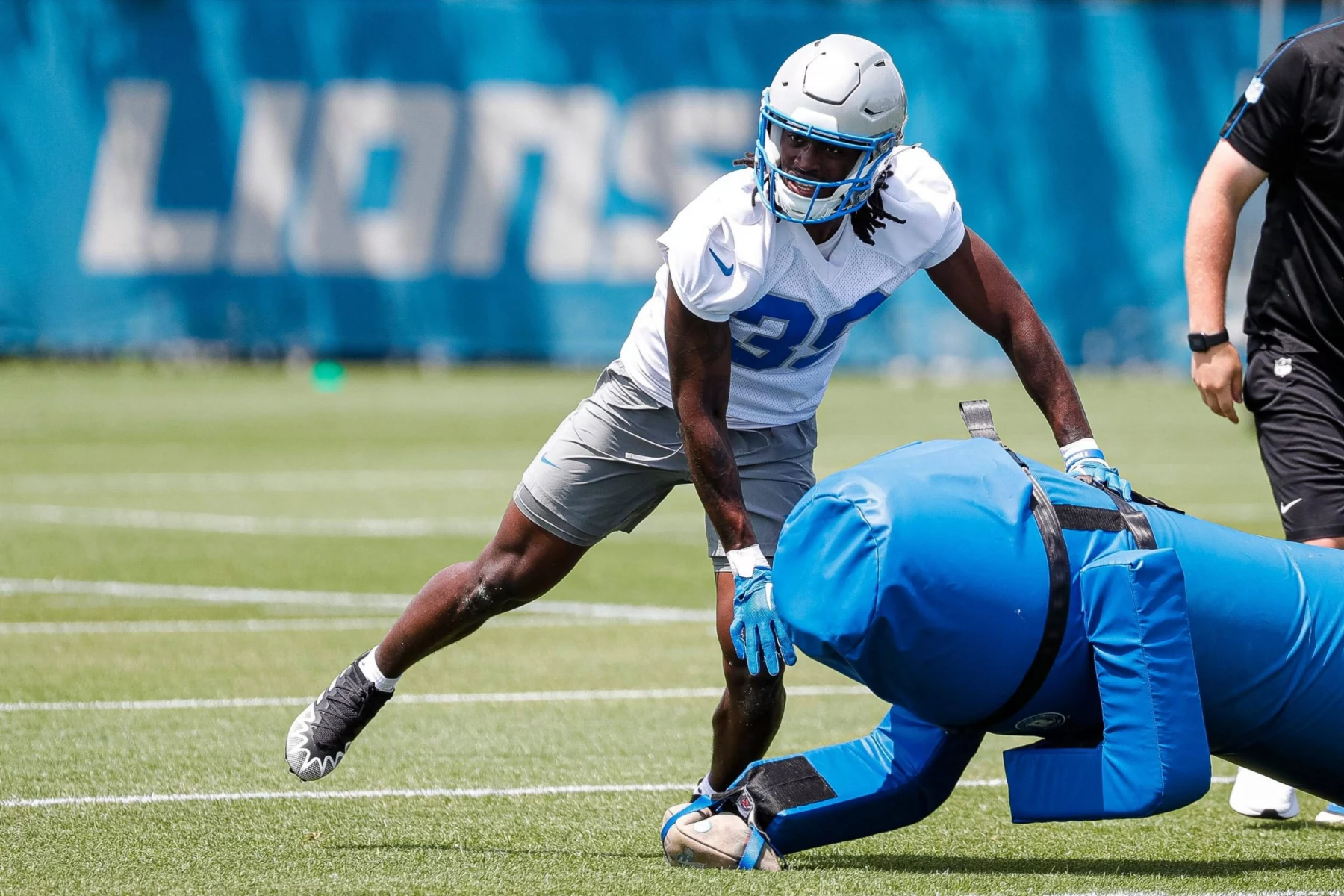 Morice Norris, ein Footballspieler in Trainingskleidung der Detroit Lions mit silbernem Helm und blauen Handschuhen, beugt sich auf dem Rasen vor, um einen blauen Tackling-Dummy zu schieben. Eine weitere Person in schwarzer Sportkleidung steht in der Nähe, im verschwommenen Hintergrund ist "LIONS" zu sehen. Diese Beschreibung wurde mit der FootballR KI automatisch generiert.