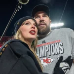 Taylor Swift mit schwarzer Mütze und Mantel steht in einem Football-Stadion neben Travis Kelce in einem grauen "Champions"-Shirt der Kansas City Chiefs und einer schwarzen Mütze, beide blicken nach vorne - vielleicht feiern sie die Verlobung des Jahres. Die Stadionbeleuchtung leuchtet hinter ihnen. Diese Beschreibung wurde mit der FootballR KI automatisch generiert.