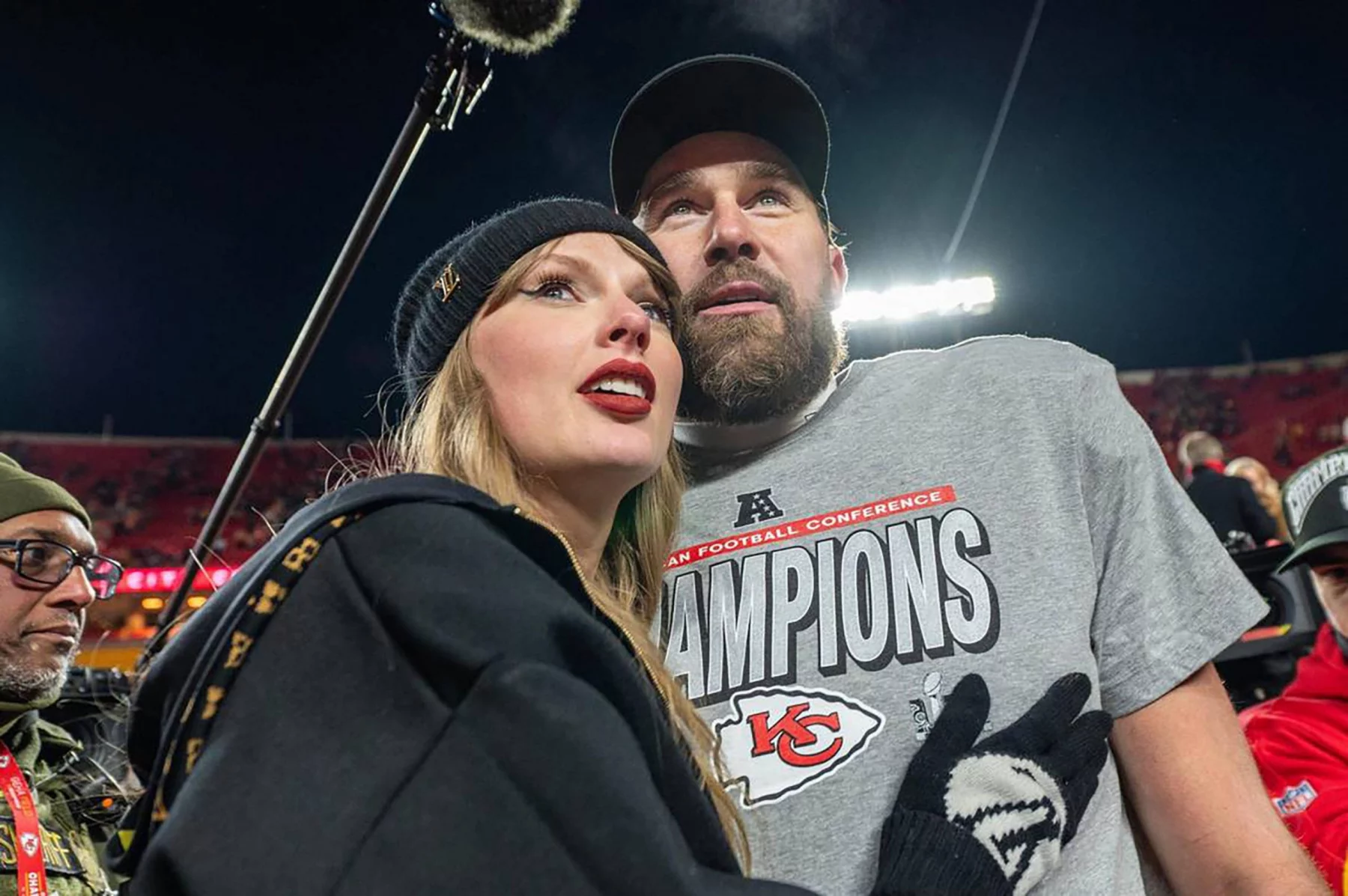 Taylor Swift mit schwarzer Mütze und Mantel steht in einem Football-Stadion neben Travis Kelce in einem grauen "Champions"-Shirt der Kansas City Chiefs und einer schwarzen Mütze, beide blicken nach vorne - vielleicht feiern sie die Verlobung des Jahres. Die Stadionbeleuchtung leuchtet hinter ihnen. Diese Beschreibung wurde mit der FootballR KI automatisch generiert.