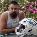 Ein muskulöser Mann mit dunklem Bart, der ein ärmelloses Miami Dolphins-Shirt trägt, lehnt sich im Freien an eine Fläche. Vor ihm steht ein weißer Football-Helm der Miami Dolphins, während Zach Sieler die Szene genießt. Rosa Blumen und Grünzeug verschwimmen im Hintergrund. Diese Beschreibung wurde mit der FootballR KI automatisch generiert.