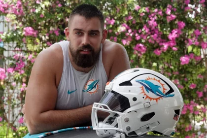 Ein muskulöser Mann mit dunklem Bart, der ein ärmelloses Miami Dolphins-Shirt trägt, lehnt sich im Freien an eine Fläche. Vor ihm steht ein weißer Football-Helm der Miami Dolphins, während Zach Sieler die Szene genießt. Rosa Blumen und Grünzeug verschwimmen im Hintergrund. Diese Beschreibung wurde mit der FootballR KI automatisch generiert.