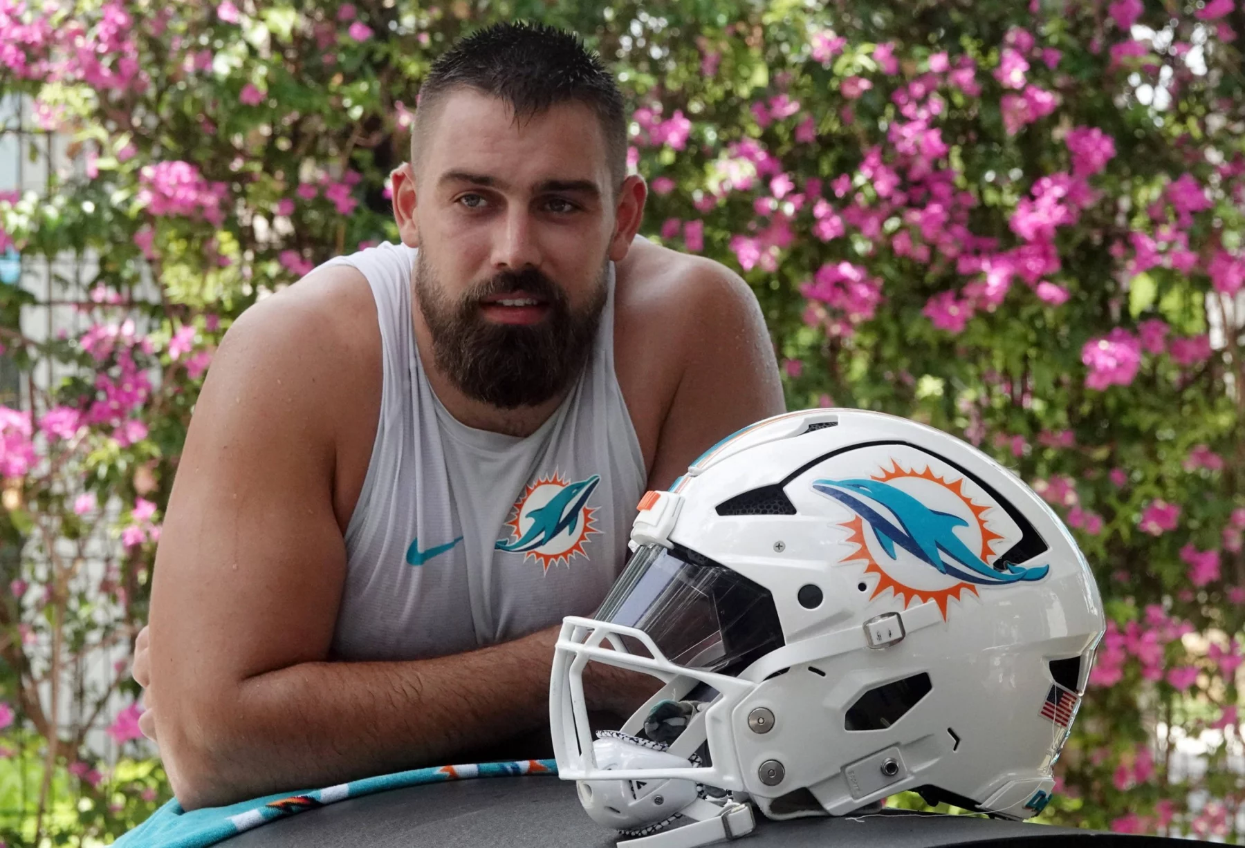 Ein muskulöser Mann mit dunklem Bart, der ein ärmelloses Miami Dolphins-Shirt trägt, lehnt sich im Freien an eine Fläche. Vor ihm steht ein weißer Football-Helm der Miami Dolphins, während Zach Sieler die Szene genießt. Rosa Blumen und Grünzeug verschwimmen im Hintergrund. Diese Beschreibung wurde mit der FootballR KI automatisch generiert.