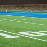 Nahaufnahme eines Football-Feldes mit weißer "10"-Yard-Linie, grünem Kunstrasen, zwei Fußbällen in der Nähe der blauen Endzone und leeren Tribünen, die die Stille vor der Rückkehr der Stars 2025 einfangen; noch sind keine Spieler zurück auf dem Feld. Diese Beschreibung wurde mit der FootballR KI automatisch generiert.