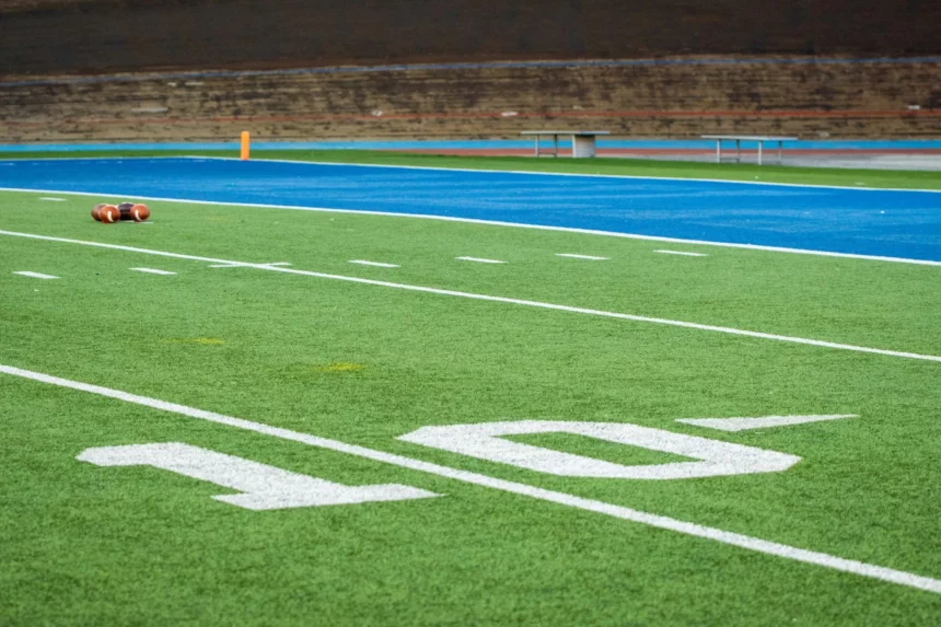 Nahaufnahme eines Football-Feldes mit weißer "10"-Yard-Linie, grünem Kunstrasen, zwei Fußbällen in der Nähe der blauen Endzone und leeren Tribünen, die die Stille vor der Rückkehr der Stars 2025 einfangen; noch sind keine Spieler zurück auf dem Feld. Diese Beschreibung wurde mit der FootballR KI automatisch generiert.