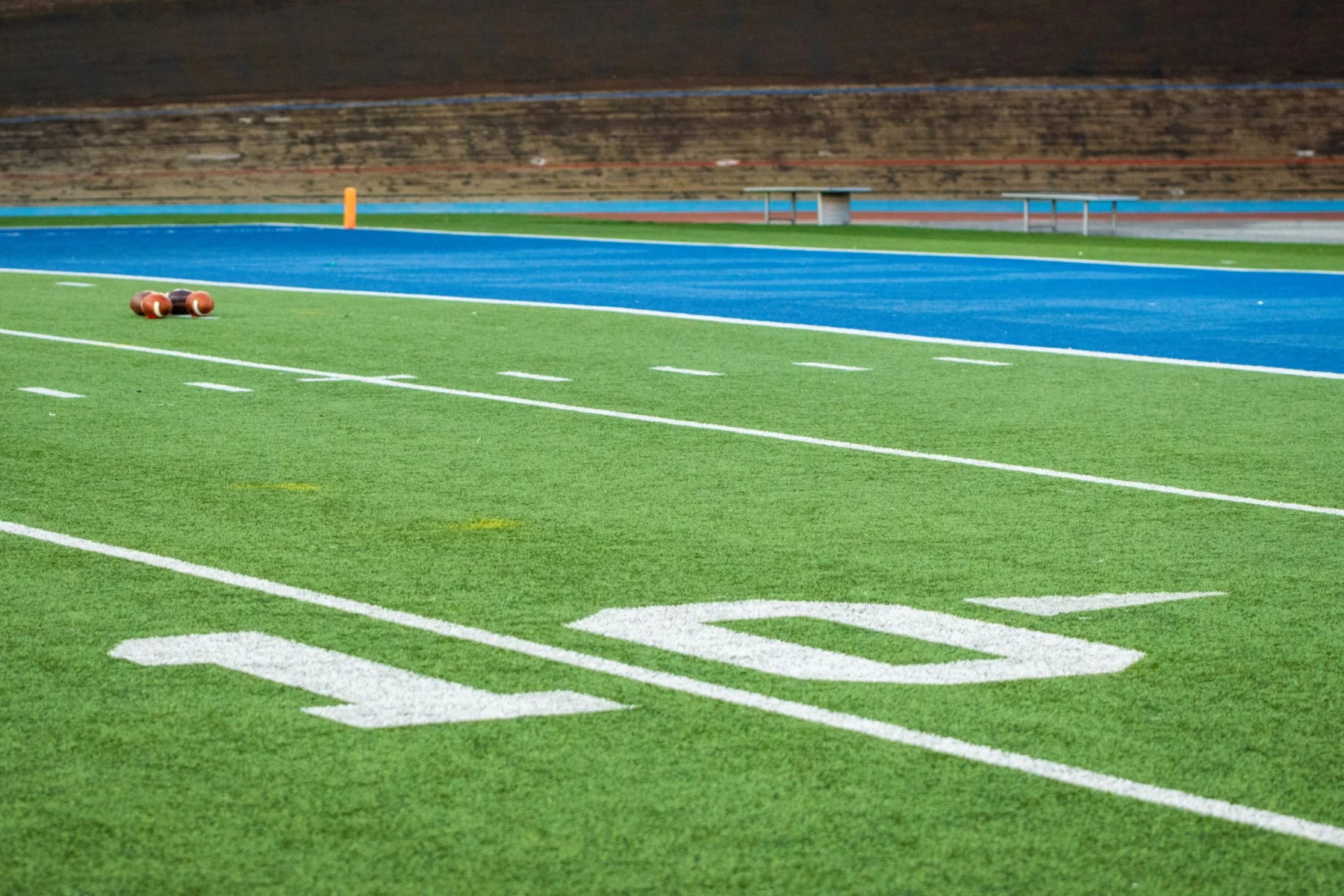 Nahaufnahme eines Football-Feldes mit weißer "10"-Yard-Linie, grünem Kunstrasen, zwei Fußbällen in der Nähe der blauen Endzone und leeren Tribünen, die die Stille vor der Rückkehr der Stars 2025 einfangen; noch sind keine Spieler zurück auf dem Feld. Diese Beschreibung wurde mit der FootballR KI automatisch generiert.