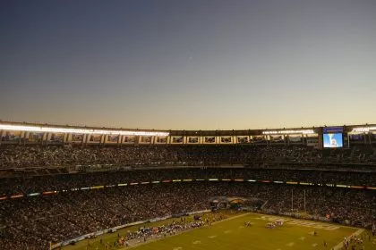Weitwinkelansicht eines überfüllten Stadions in der Abenddämmerung während der NFL-Saison. Das Spielfeld ist hell erleuchtet und die Fans füllen die Tribünen. Der klare Himmel zeigt eine schwache Mondsichel, während eine große Leinwand eine Nahaufnahme zeigt und Banner den Oberrang schmücken. Diese Beschreibung wurde mit der FootballR KI automatisch generiert.