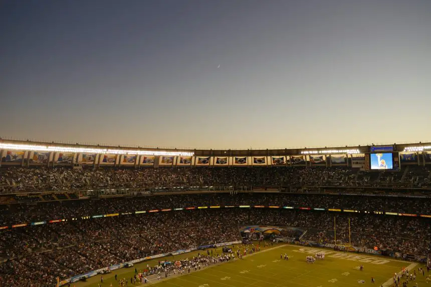 Weitwinkelansicht eines überfüllten Stadions in der Abenddämmerung während der NFL-Saison. Das Spielfeld ist hell erleuchtet und die Fans füllen die Tribünen. Der klare Himmel zeigt eine schwache Mondsichel, während eine große Leinwand eine Nahaufnahme zeigt und Banner den Oberrang schmücken. Diese Beschreibung wurde mit der FootballR KI automatisch generiert.