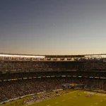 Weitwinkelansicht eines überfüllten Stadions in der Abenddämmerung während der NFL-Saison. Das Spielfeld ist hell erleuchtet und die Fans füllen die Tribünen. Der klare Himmel zeigt eine schwache Mondsichel, während eine große Leinwand eine Nahaufnahme zeigt und Banner den Oberrang schmücken. Diese Beschreibung wurde mit der FootballR KI automatisch generiert.