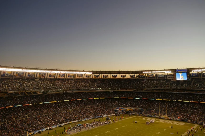 Weitwinkelansicht eines überfüllten Stadions in der Abenddämmerung während der NFL-Saison. Das Spielfeld ist hell erleuchtet und die Fans füllen die Tribünen. Der klare Himmel zeigt eine schwache Mondsichel, während eine große Leinwand eine Nahaufnahme zeigt und Banner den Oberrang schmücken. Diese Beschreibung wurde mit der FootballR KI automatisch generiert.