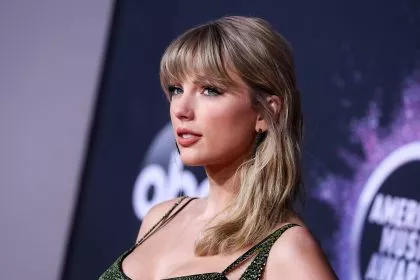 Taylor Swift mit langen blonden Haaren und Pony, posiert in einem glitzernden grünen Kleid auf einem roten Teppich vor einem dunklen Super Bowl LX-Hintergrund mit unscharfen kreisförmigen Logos, der leicht zur Seite blickt und einen gelassenen Ausdruck hat. Diese Beschreibung wurde mit der FootballR KI automatisch generiert.