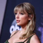 Taylor Swift mit langen blonden Haaren und Pony, posiert in einem glitzernden grünen Kleid auf einem roten Teppich vor einem dunklen Super Bowl LX-Hintergrund mit unscharfen kreisförmigen Logos, der leicht zur Seite blickt und einen gelassenen Ausdruck hat. Diese Beschreibung wurde mit der FootballR KI automatisch generiert.