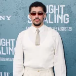 Bad Bunny - Ein Mann mit kurzen lockigen Haaren, Spitzbart und Sonnenbrille steht vor einem blauen "CAUGHT STEALING"-Hintergrund bei einem Sony-Event. Er ist weiß gekleidet und verkörpert den kultigen Stil von Bad Bunny - perfekt für eine Halftime Show oder einen Auftritt beim Super Bowl. Diese Beschreibung wurde mit der FootballR KI automatisch generiert.