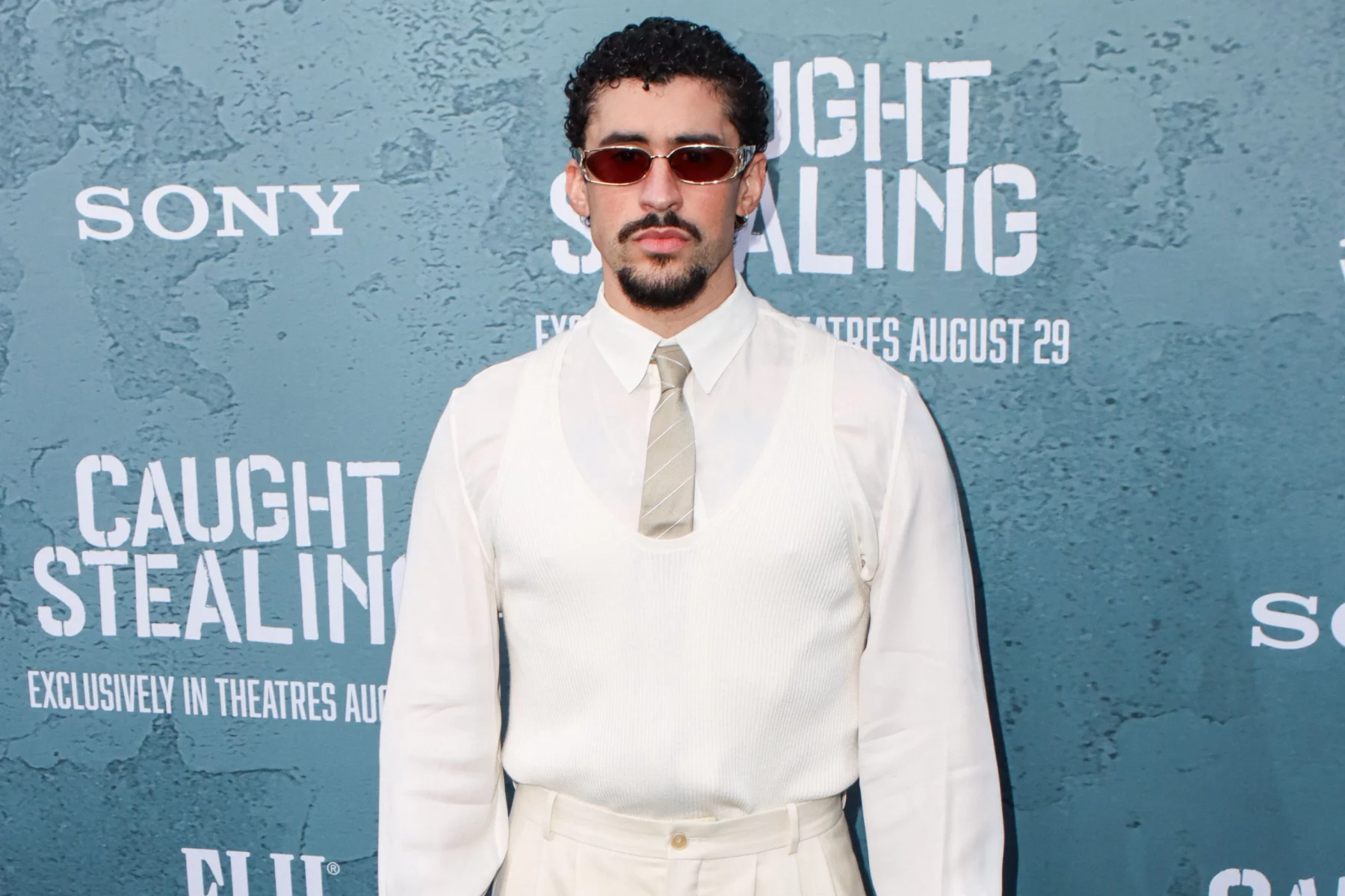 Bad Bunny - Ein Mann mit kurzen lockigen Haaren, Spitzbart und Sonnenbrille steht vor einem blauen "CAUGHT STEALING"-Hintergrund bei einem Sony-Event. Er ist weiß gekleidet und verkörpert den kultigen Stil von Bad Bunny - perfekt für eine Halftime Show oder einen Auftritt beim Super Bowl. Diese Beschreibung wurde mit der FootballR KI automatisch generiert.