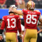 Zwei Footballspieler der San Francisco 49ers laufen Seite an Seite in Rot und Gold. Die Nummer 13, Brock Purdy, schaut weg, während die Nummer 85, George Kittle, eine Hand liebevoll auf seinen Helm legt - ein seltener lichter Moment inmitten der Verletzungsmisere der 49ers. Der Hintergrund ist unscharf. Diese Beschreibung wurde mit der FootballR KI automatisch generiert.
