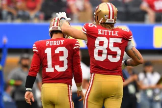 Woche 3 - Zwei Footballspieler der San Francisco 49ers laufen Seite an Seite in Rot und Gold. Die Nummer 13, Brock Purdy, schaut weg, während die Nummer 85, George Kittle, eine Hand liebevoll auf seinen Helm legt - ein seltener lichter Moment inmitten der Verletzungsmisere der 49ers. Der Hintergrund ist unscharf. Diese Beschreibung wurde mit der FootballR KI automatisch generiert.