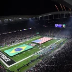 Rio de Janeiro - Ein volles Football-Stadion bei Nacht, in dem ein Spiel der NFL International Series stattfindet, mit großen brasilianischen und amerikanischen Flaggen auf dem Spielfeld. Feuerwerkskörper erhellen den Himmel, während "EAGLES" die Endzone markiert, und Zuschauer aus Brasilien füllen die beleuchteten Tribünen. Diese Beschreibung wurde mit der FootballR KI automatisch generiert.