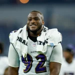 Nnamdi Madubuike in einem Baltimore-Ravens-Trikot mit der Nummer 92 steht auf dem Spielfeld. Er hat sich ein weißes Handtuch um den Hals gewickelt, möglicherweise nachdem er sich von einer Nackenverletzung erholt hat. Im Hintergrund des Stadions sind weitere Personen zu sehen. Diese Beschreibung wurde mit der FootballR KI automatisch generiert.