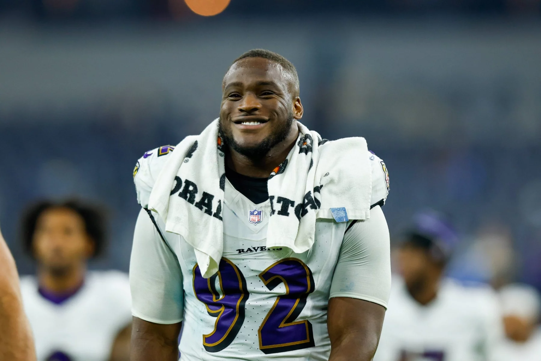 Nnamdi Madubuike in einem Baltimore-Ravens-Trikot mit der Nummer 92 steht auf dem Spielfeld. Er hat sich ein weißes Handtuch um den Hals gewickelt, möglicherweise nachdem er sich von einer Nackenverletzung erholt hat. Im Hintergrund des Stadions sind weitere Personen zu sehen. Diese Beschreibung wurde mit der FootballR KI automatisch generiert.