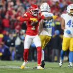 Ein Football-Spieler der Kansas City Chiefs in einer roten Uniform zeigt auf einen Football und feiert auf dem Spielfeld. Xavier Worthy, in einem weiß-gelben Chargers-Trikot mit der Nummer 25, steht in der Nähe, während ein anderer Chargers-Spieler und Fans im Hintergrund jubeln. Diese Beschreibung wurde mit der FootballR KI automatisch generiert.