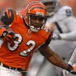 Ein Footballspieler der Cincinnati Bengals, Rudi Johnson, trägt ein orangefarbenes Trikot mit der Nummer 32. Er läuft mit dem Ball und weicht Verteidigern in Weiß und Silber aus. Der Ex-Bengals-Star rennt über das Feld, während im Hintergrund unscharfe Spieler zu sehen sind. Diese Beschreibung wurde mit der FootballR KI automatisch generiert.